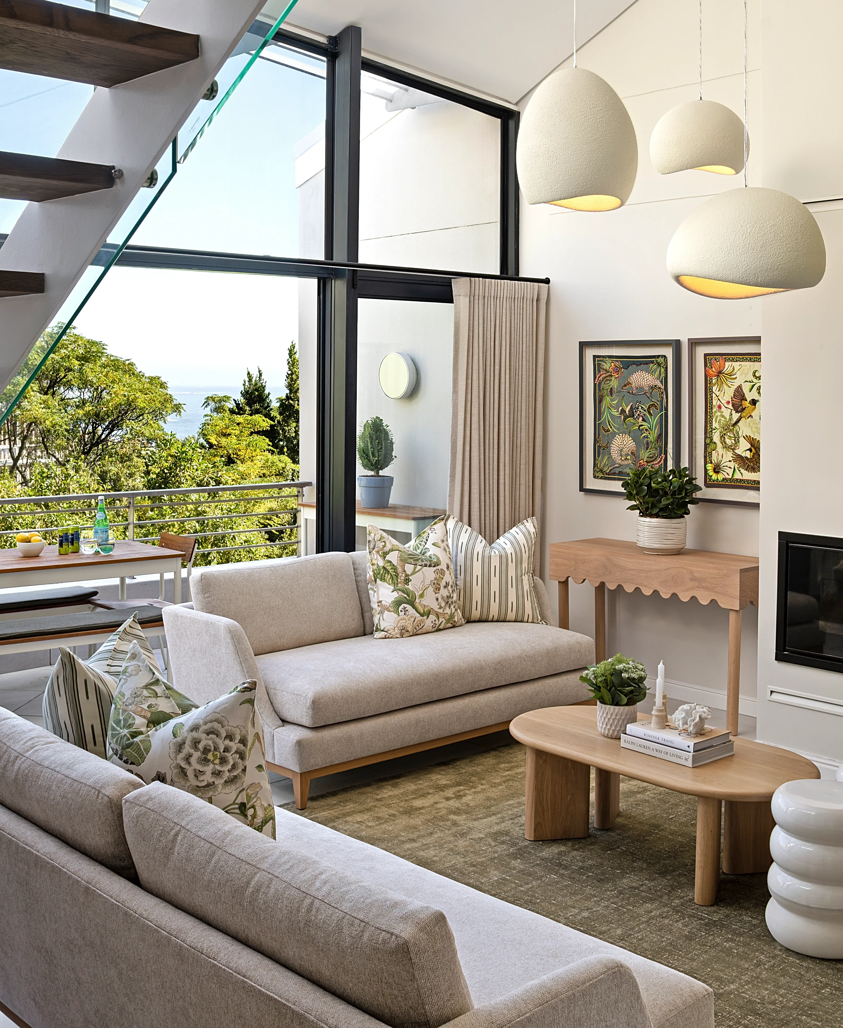 Living room at The Glen, Camps Bay: three cream pendant lights, a cream sofa and loveseat with floral and striped cushions, an oak coffee table, framed botanical prints and black-framed windows onto the trees