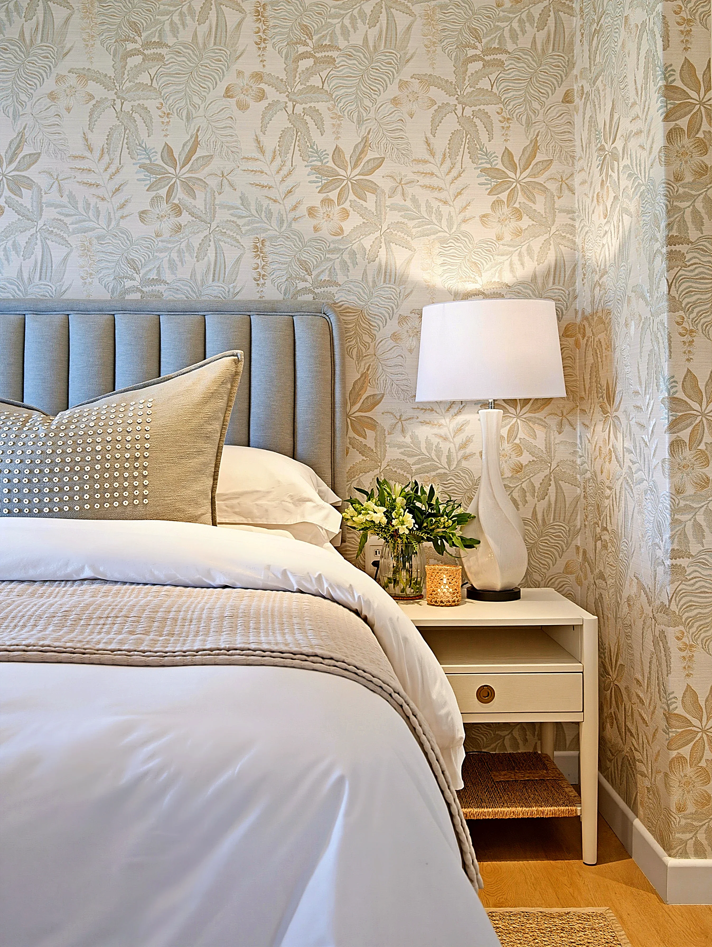 Bedroom at Simbithi Eco-Estate: botanical wallpaper, a grey-blue channel-stitched headboard, a beaded cushion, a white lamp and a cream bedside table